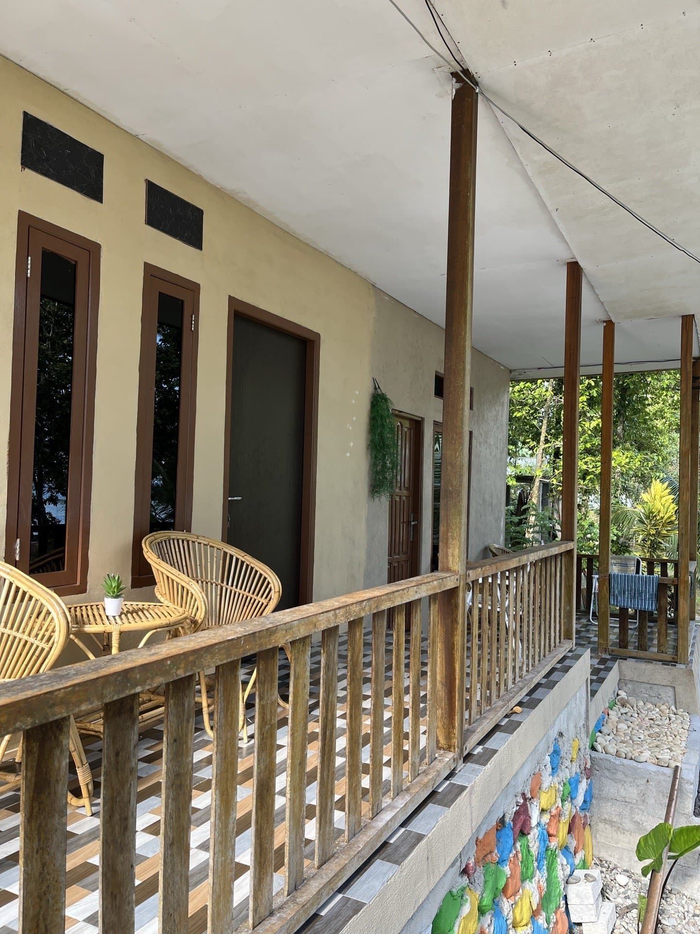 Teras kamar Shafira Resort dengan kursi rotan, jendela berbingkai kayu cokelat, dan taman batu warna-warni di Pantai Memit Morella